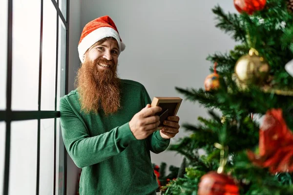 Young Redhead Man Looking Photo Standing Christmas Tree Home — Φωτογραφία Αρχείου