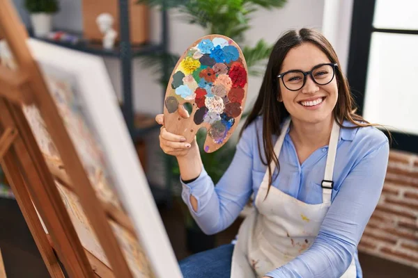 年轻的黑发女子牵着画家的调色板 站在艺术工作室里 面带微笑 面带微笑 面带微笑 显得积极而快乐 — 图库照片