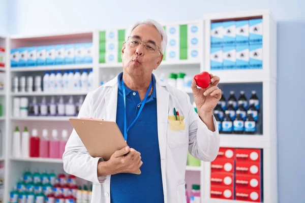 Middle age man with grey hair working at pharmacy drugstore holding red heart making fish face with mouth and squinting eyes, crazy and comical.
