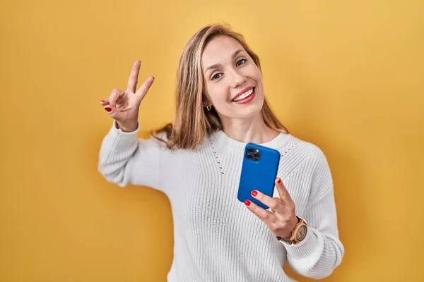 スマートフォンの入力メッセージを使用して若いブロンドの女性は勝利のサインを行う指を示すカメラを見て笑顔 — ストック写真