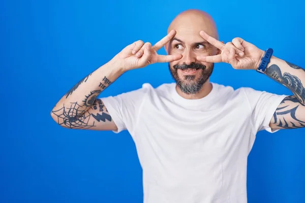 有纹身的西班牙裔男子站在蓝色的背景上 用手指捂住脸做和平的象征 笑容满面地表示胜利 — 图库照片