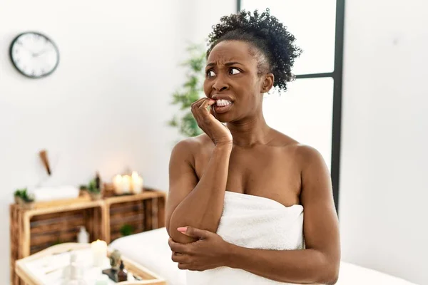 Young african american woman wearing towel standing at beauty center looking stressed and nervous with hands on mouth biting nails. anxiety problem.