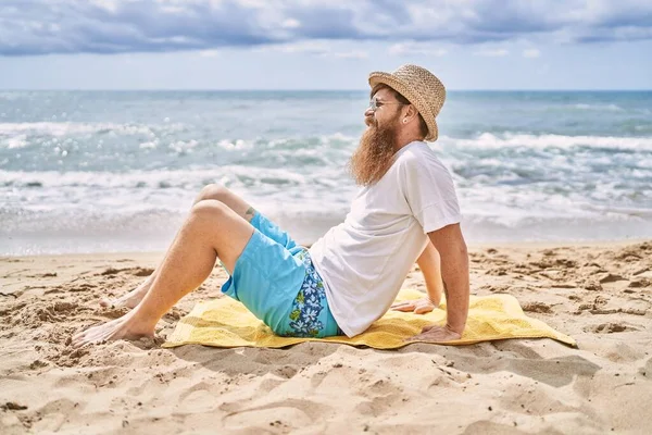 若い赤毛の男は ビーチでタオルの上に座って幸せな笑顔 — ストック写真