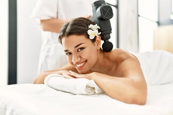 Young latin woman relaxed having back massage with percussion pistol at beauty center
