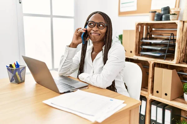 Black woman with braids working at the office speaking on the phone smiling looking to the side and staring away thinking.