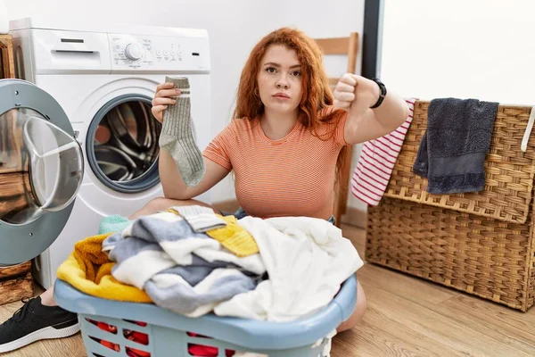 若い赤毛の女性は 洗濯機に汚れた洗濯機を入れますが 親指を下にジェスチャーで拒絶と否定を示す不満と怒りに見えます 悪い表現 — ストック写真