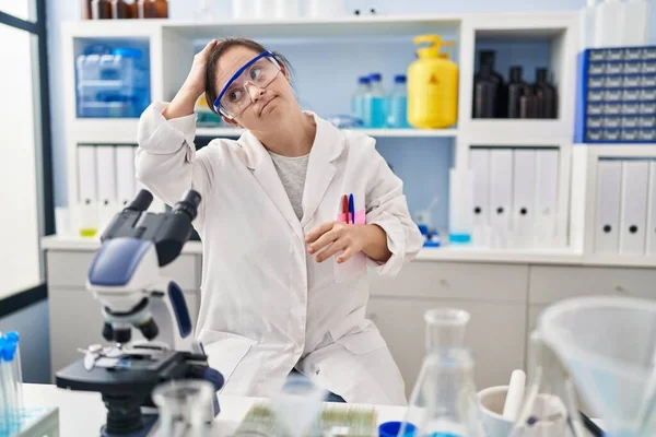 Hispanic girl with down syndrome working at scientist laboratory confuse and wondering about question. uncertain with doubt, thinking with hand on head. pensive concept.