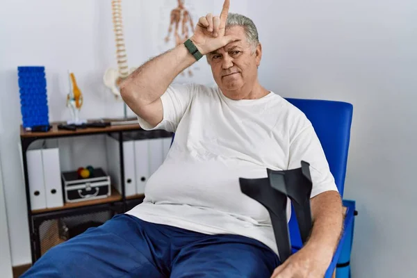 Homem Caucasiano Sênior Clínica Fisioterapia Segurando Muletas Zombando Pessoas Com — Fotografia de Stock