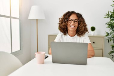 Orta yaşlı İspanyol kadın evde dizüstü bilgisayar kullanarak gülümsüyor.