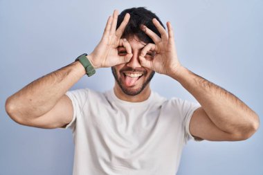 Beyaz arka planda sakallı İspanyol bir adam dürbün gibi jest yapıyor, gözleri parmaklarından bakıyor. çılgın ifade. 