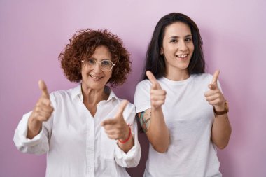 İspanyol anne ve kızı pembe arka plan üzerine gündelik beyaz tişört giyip mutlu ve komik yüzlü parmakları kameraya doğrultuyorlar. iyi enerji ve titreşimler. 