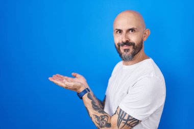 Mavi arka planda dikilen İspanyol dövmeli adam avuçları açık, fotokopi alanını gösteren, gülümseyerek mutlu bir gülümseme sunan. 