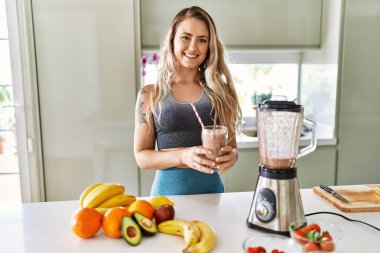 Mutfakta elinde bir bardak meyve püresi tutarak gülümseyen genç bir kadın.
