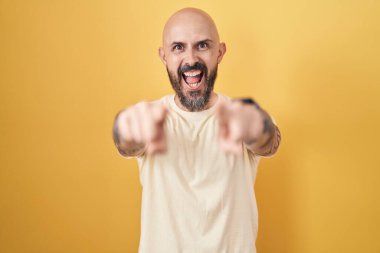 Sarı arka planda dikilen İspanyol dövmeli adam seni ve kamerayı işaret ediyor, gülümsüyor olumlu ve neşeli. 