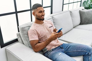 Genç Arap adam evde akıllı telefon kullanarak kendinden emin gülümsüyor.