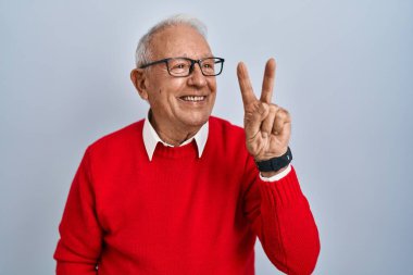 İzole edilmiş arka planda gri saçlı yaşlı adam kameraya bakıp zafer işareti yaparken gülümsüyor. İki numara.. 