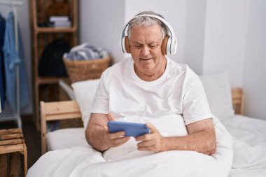 Orta yaşlı, kır saçlı, video oyunu oynayan, yatak odasında oturan bir adam.