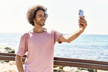 Mutlu gülümseyen genç İspanyol adam sahilde duran akıllı telefonun yanında selfie çekiyor..