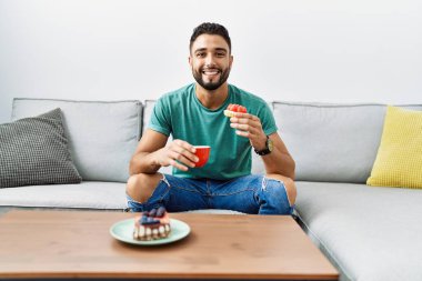 Genç Arap adam kahvaltısını evde, kanepede yapıyor.