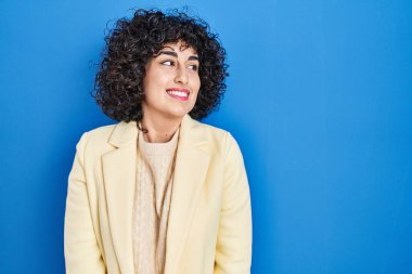 Kıvırcık saçlı genç esmer kadın mavi arka planda duruyor gülümsüyor ve yan tarafa bakıyor ve düşünüyor.. 