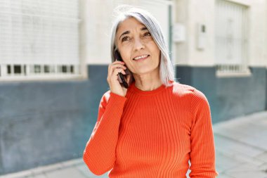 Orta yaşlı, kır saçlı, gülümseyen, mutlu, akıllı telefondan konuşan bir kadın..