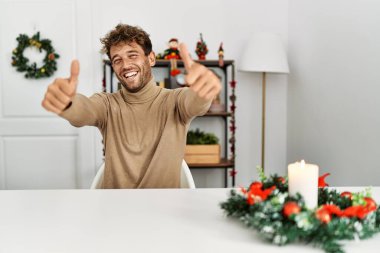 Yılbaşı süslemelerine kadar masanın üzerinde oturan sakallı yakışıklı genç adam elleriyle olumlu bir jest yapmayı onaylıyor, gülümsüyor ve başarı için seviniyor. kazanan jest. 