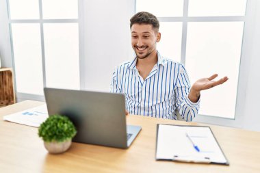 Ofiste çalışan sakallı yakışıklı bir genç. Bilgisayarı kullanıyor. Gülümsüyor, gülümsüyor ve avuç içi kameraya bakıyor.. 