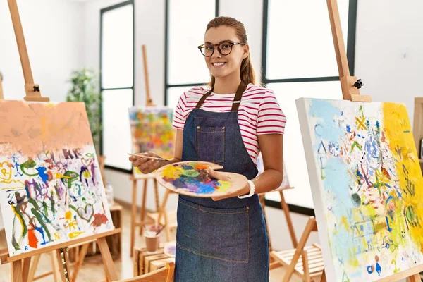 年轻女子笑着自信地拿着画笔和调色板在艺术工作室里 — 图库照片