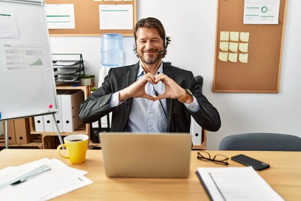 Handsome middle age man wearing call center agent headset at the office smiling in love doing heart symbol shape with hands. romantic concept.