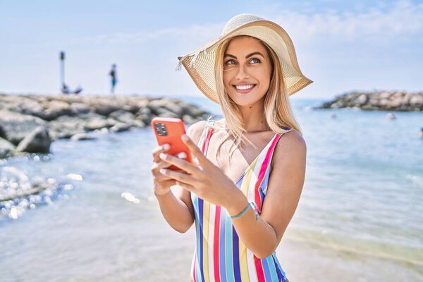 Young blonde girl wearing summer hat using smartphone at the beach.