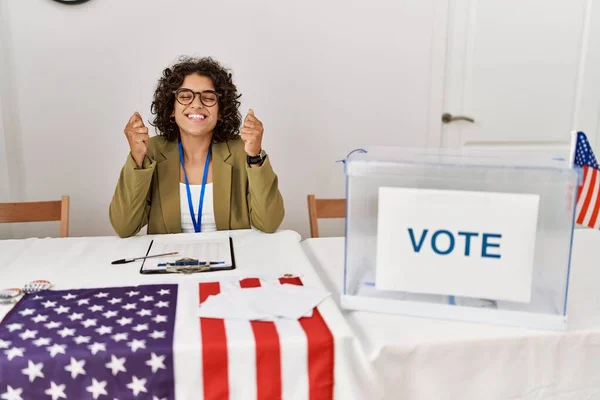 Young hispanic woman at political election sitting by ballot excited for success with arms raised and eyes closed celebrating victory smiling. winner concept.