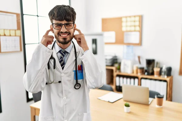 Hispanic man with beard wearing doctor uniform and stethoscope at the office covering ears with fingers with annoyed expression for the noise of loud music. deaf concept.