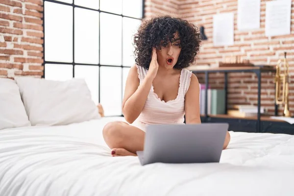 若い中東の女性はベッドの上でコンピュータのラップトップを使用して恐怖と驚きのために口を開けて驚いて 不信の顔 — ストック写真