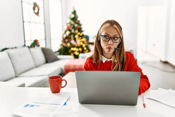 年轻的高加索女孩坐在桌子上 用笔记本电脑在圣诞树旁工作 用嘴唇 疯狂和滑稽的姿势做着鱼脸 有趣的表达方式 — 图库照片