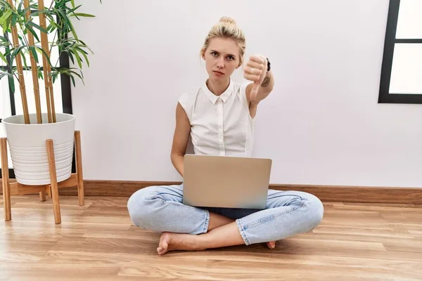 Young blonde woman using computer laptop sitting on the floor at the living room looking unhappy and angry showing rejection and negative with thumbs down gesture. bad expression.