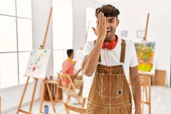 Young hispanic man at art studio covering one eye with hand, confident smile on face and surprise emotion.