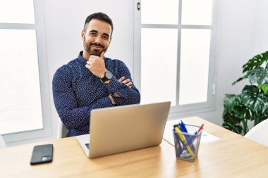 Ofiste sakallı, laptopuyla çalışan, kollarını kavuşturup el pençe divan duran ve kameraya gülümseyen genç İspanyol bir adam. Olumlu düşünmek. 