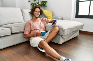 Genç İspanyol adam kahve içiyor ve akıllı telefon kullanıyor. Evde yerde oturuyor..