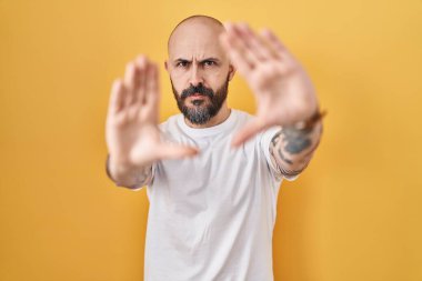 Sarı arkaplanda dikilen dövmeli genç İspanyol adam el ve parmaklarıyla çerçeve yapıyor, kamera açısı var. 