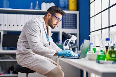 Young hispanic man wearing scientist uniform using laptop and microscope at laboratory