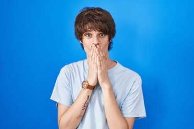 Mavi arka planda dikilen İspanyol genç adam gülüyor ve utanıyor kıkır kıkır kıkır kıkır gülüyor elleri, dedikoduları ve skandalları anlatıyor 