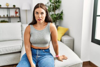 Genç esmer kadın evde şaşkın bir şekilde kanepede oturuyor, şüpheci ve alaycı bakıyor, ağzı açık kalmış. 