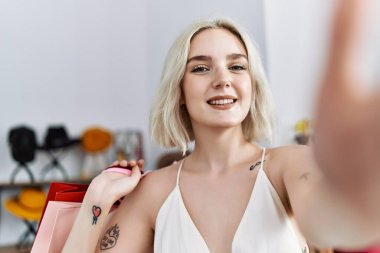 Young caucasian woman holding shopping bags make selfie by the camera at clothing store