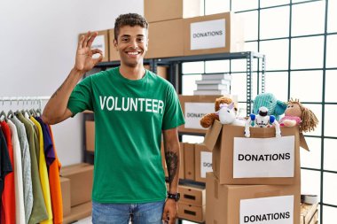 Bağışlarda gönüllü tişört giyen yakışıklı İspanyol genç adam gülümseyerek el ve parmaklarıyla 