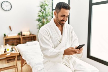 Genç İspanyol bornoz giyen adam güzellik merkezinde akıllı telefon kullanıyor.