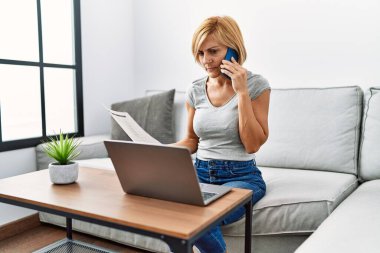 Orta yaşlı sarışın kadın evde dizüstü bilgisayar kullanarak akıllı telefondan konuşuyor.