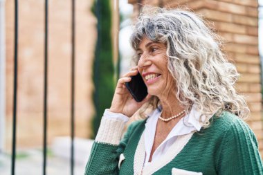 Orta yaşlı bir kadın gülümsüyor. Sokakta akıllı telefondan konuşuyor.