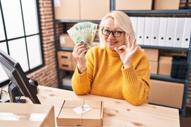 Orta yaşlı beyaz bir kadın küçük işletme şirketinde çalışıyor elinde Hong Kong dolarları var tamam imzasını atıyor parmaklarıyla gülümseyerek mükemmel bir sembol oluşturuyor. 