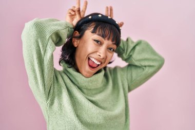 Young beautiful woman standing over pink background posing funny and crazy with fingers on head as bunny ears, smiling cheerful 