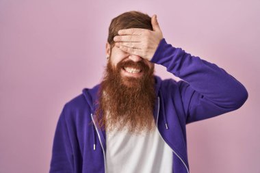 Pembe arka planda uzun sakallı beyaz bir adam. Gülümsüyor ve gülümsüyor. Gözleri şaşkınlıkla örtüyor. kör kavram. 